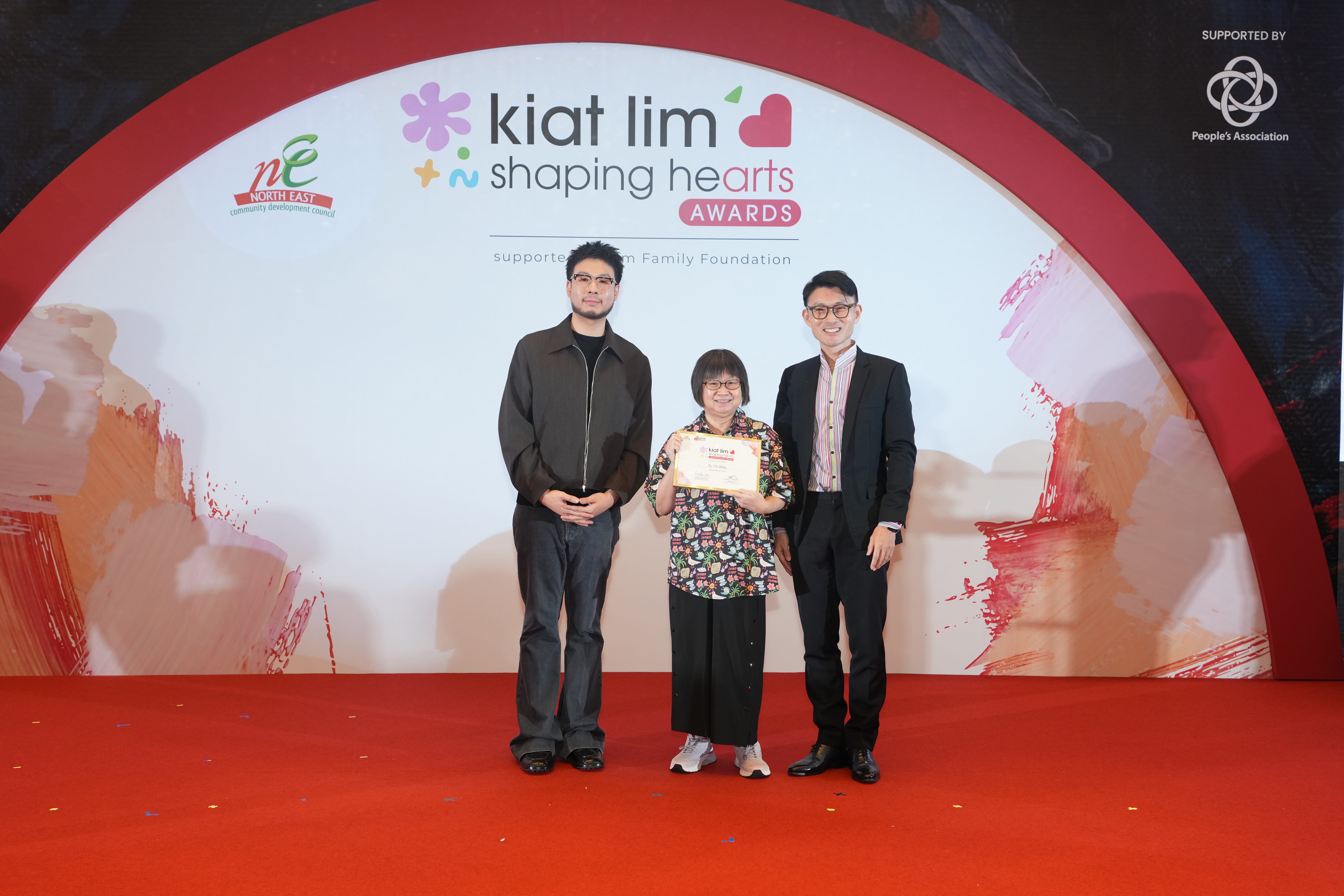 Award recipient standing on stage with the presenter, holding their certificate and posing for a group photo during the award ceremony, with a backdrop reading ‘Kiat Lim-Shaping Hearts Awards'.